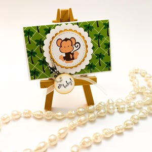 May include: A green and white card with a cartoon monkey on it, sitting on a small wooden easel. The card has a white circle with a gold border around the monkey. The card also has a small wooden tag with the text "Oh baby!" hanging from it.