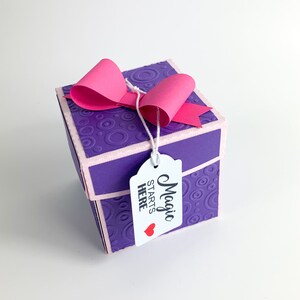 May include: A purple gift box with a pink bow and a white tag that says "Magic Starts Here" with a red heart.