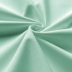 May include: A close-up of a soft, smooth, light green fabric with a subtle texture. The fabric is folded and draped, showcasing its draping qualities.