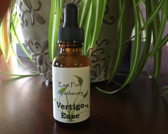 May include: A brown glass bottle with a black dropper, labeled "East Fork Apothecary Vertigo Ease." The bottle is set against a backdrop of green and white leafy plants and a decorative gray pot.