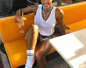 May include: A woman wearing a white tank top, denim shorts, white sneakers with colorful paint splatters, and a silver chain necklace sits on a yellow couch with a cocktail in her hand.