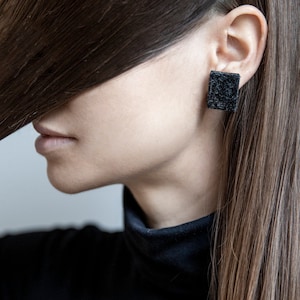 May include: A woman with long brown hair wearing black square earrings. The earrings are made of a textured material.