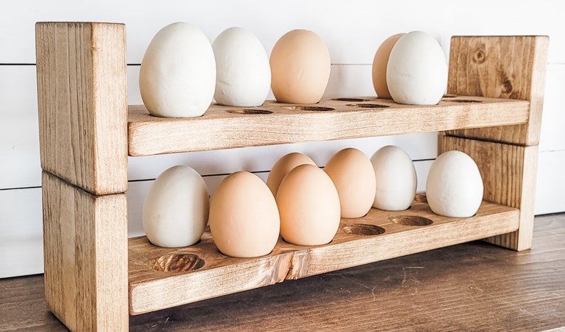 May include: A two-tiered wooden egg holder with a rustic finish. The holder is filled with a dozen eggs, some white and some light brown. The design features a series of circular cutouts to hold the eggs securely. The wooden structure is sturdy and well-crafted.