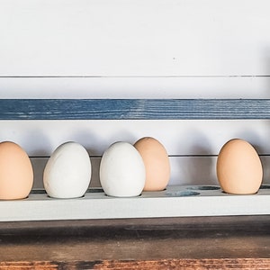 May include: Wooden egg holder painted in shades of blue and white, holding six eggs. The eggs are a mix of white and light brown. The holder has a rustic, farmhouse style.