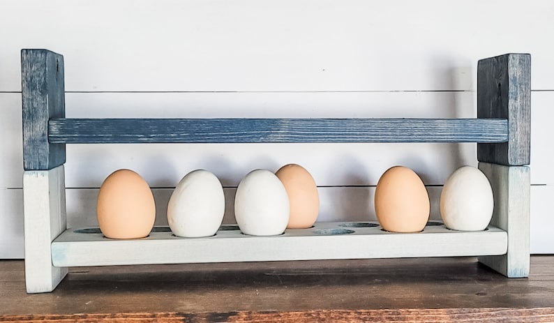May include: Wooden egg holder painted in shades of blue and white, holding six eggs. The eggs are a mix of white and light brown. The holder has a rustic, farmhouse style.