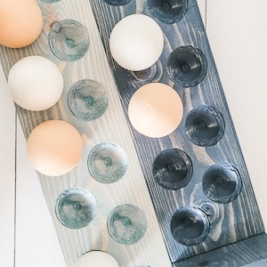 May include: Two wooden egg holders, one light blue and one dark blue, each with carved egg-shaped depressions. The holders are filled with a mix of white and tan eggs. The wooden holders are on a white surface.