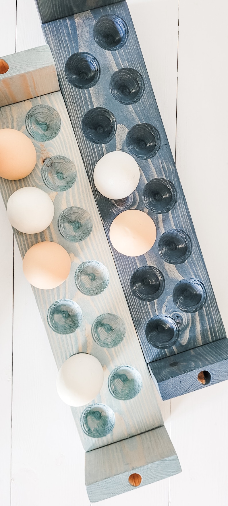 May include: Two wooden egg holders, one light blue and one dark blue, each with carved egg-shaped depressions. The holders are filled with a mix of white and tan eggs. The wooden holders are on a white surface.
