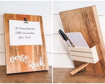 Könnte beinhalten: Holz-Klemmbrett mit Metallclip, der ein weißes Papier mit dem Text "As I have loved you, LOVE one another John 13:34" hält. Die Worte "verse of the week" sind in weißer Schrift geschrieben. Ein kleiner Holzhalter enthält einen Notizblock und einen schwarzen Marker.