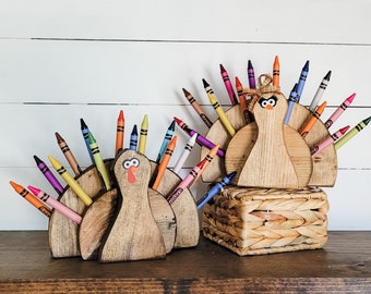 Puede incluir: Tres porta-crayones de madera con forma de pavo con crayones de colores dispuestos en forma de abanico. Los pavos tienen pintura marrón y negra y ojos saltones. Los crayones están dispuestos en forma de abanico detrás de los pavos. Los pavos están sentados sobre una superficie de madera.