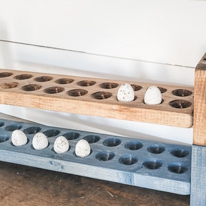 May include: A two-tiered wooden egg holder with a natural wood top and a blue-painted bottom. The holder has multiple circular openings to hold eggs. Several speckled eggs are placed in the openings, showcasing a rustic farmhouse style.
