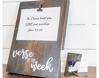 Könnte beinhalten: Ein dunkelbraunes Holzschild mit einer weißen Aufschrift, die "verse of the week" lautet. Ein weißes Papier mit einem schwarzen Clip ist an dem Schild befestigt und enthält den Text "As I have loved you, LOVE one another John 13:34".