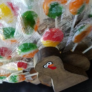 May include: A wooden turkey figurine with a painted face and red wattle, holding a variety of colorful lollipop candies wrapped in clear plastic.