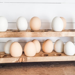 May include: A two-tiered, rustic wooden egg holder with a natural finish. The holder is filled with a mix of white and brown eggs. The design features a series of circular cutouts to hold the eggs securely. The background is a white-painted wood.