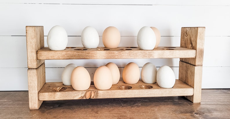 May include: A two-tiered, rustic wooden egg holder with a natural finish. The holder is filled with a mix of white and brown eggs. The design features a series of circular cutouts to hold the eggs securely. The background is a white-painted wood.
