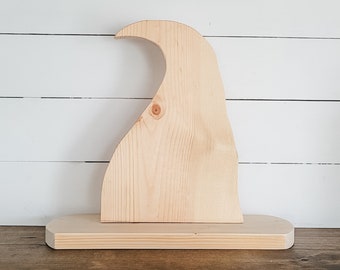 May include: A light-colored, unfinished wooden cutout shaped like a curved, pointed hat or wave, mounted vertically onto a flat, rectangular wooden base. The natural wood grain is visible throughout the piece, featuring a small knot on the front.