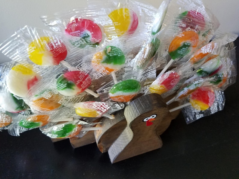 May include: A wooden turkey figurine with a red beak and eyes holds a variety of colorful lollipop candies wrapped in clear plastic.