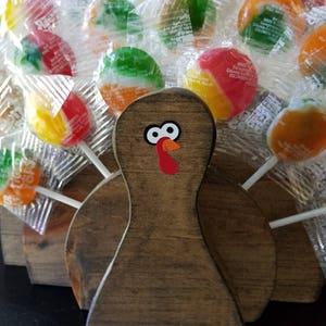 May include: A wooden turkey figurine with a red and white turkey head and eyes. The turkey is surrounded by colorful round lollipops wrapped in clear plastic.