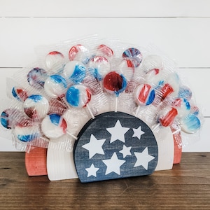 May include: A wooden stand shaped like a half-circle with white and red stripes and a black section with white stars. The stand is filled with red, white, and blue lollipops.