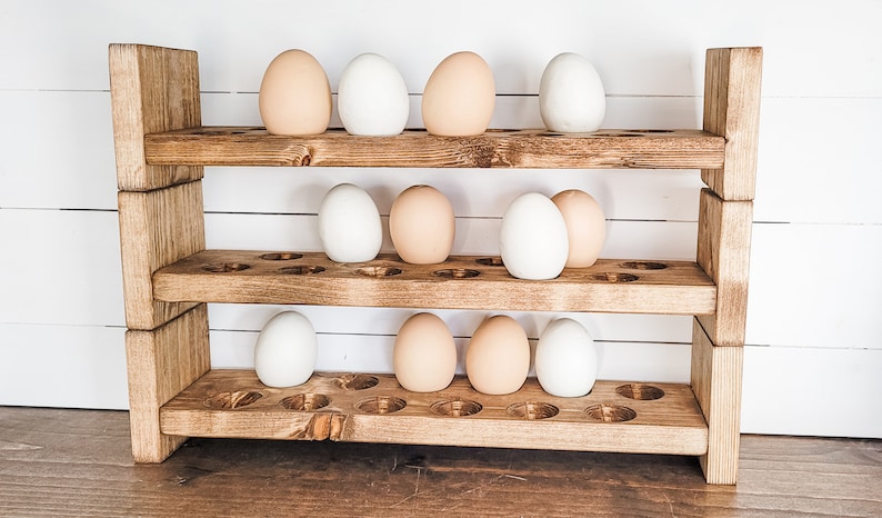 May include: A three-tiered wooden egg holder with a rustic finish. The holder is filled with a mix of white and light brown eggs. The wooden structure has a natural, warm tone, and the design is simple and functional.