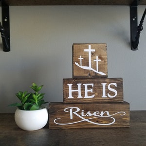 May include: Wooden block sign with the words "He is Risen" and three white crosses on a brown background.