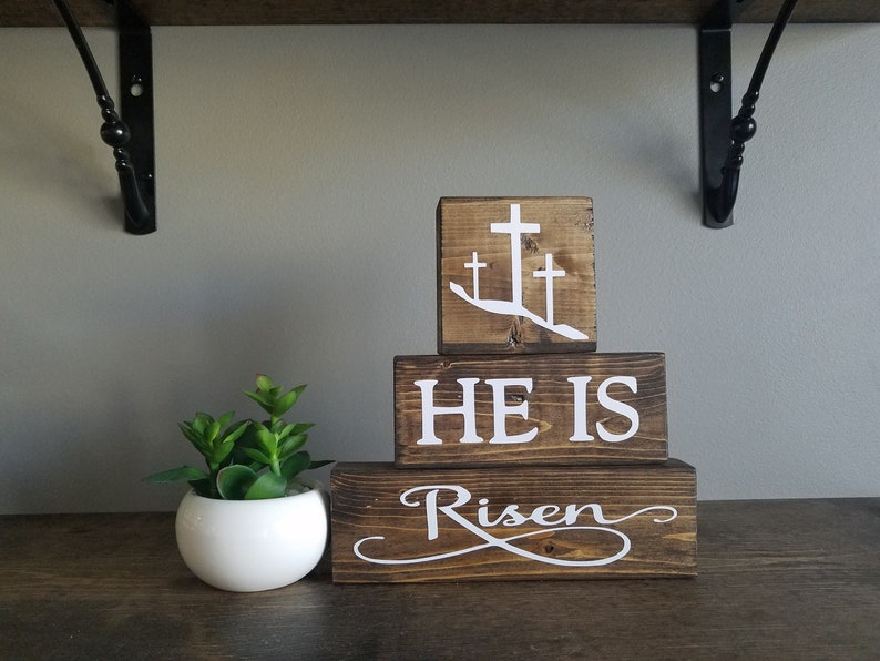 May include: Wooden block sign with the words "He is Risen" and three white crosses on a brown background.