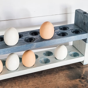 May include: A two-tiered wooden egg holder painted in shades of blue and white. The holder has circular cutouts to hold eggs. Several eggs, some white and some brown, are placed in the holder. The wooden structure is set against a white wall.