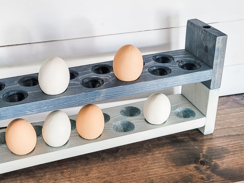 May include: A two-tiered wooden egg holder painted in shades of blue and white. The holder has circular cutouts to hold eggs. Several eggs, some white and some brown, are placed in the holder. The wooden structure is set against a white wall.