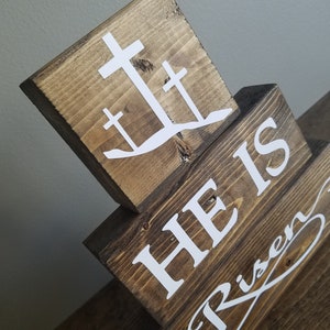 May include: Wooden block sign with the words "He is Risen" in white lettering. The top block features a white silhouette of three crosses.