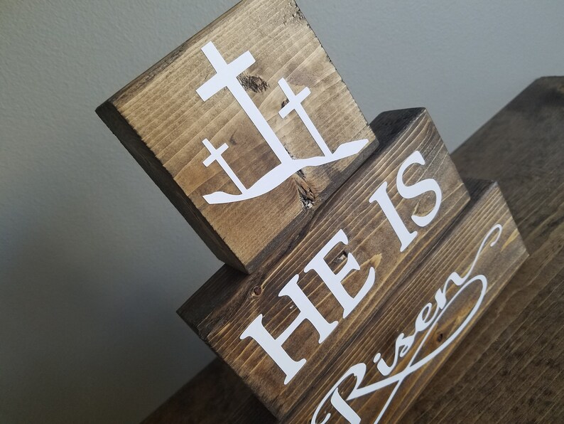 May include: Wooden block sign with the words "He is Risen" in white lettering. The top block features a white silhouette of three crosses.