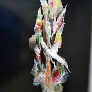 May include: A collection of colorful lollipops wrapped in clear plastic, attached to a wooden dowel. The lollipops are arranged in a circular pattern, with the sticks pointing outwards.