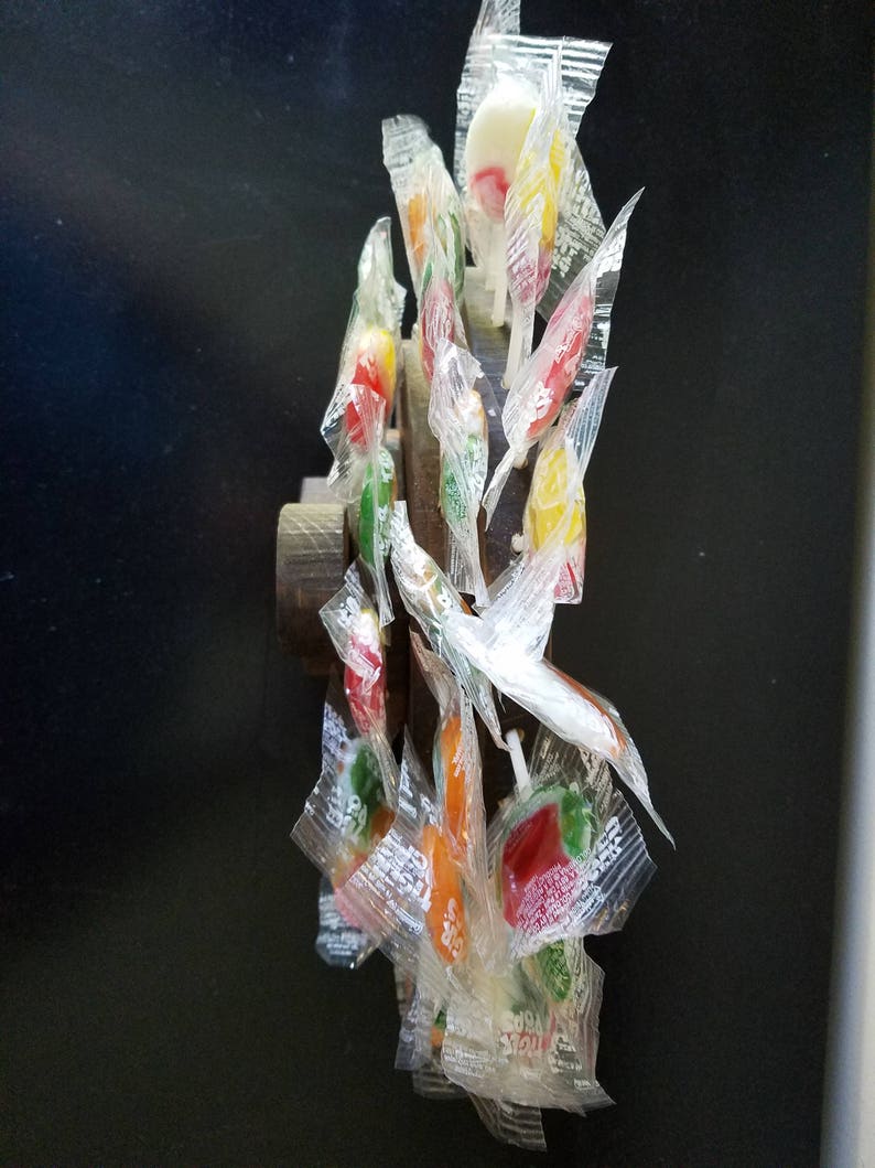 May include: A collection of colorful lollipops wrapped in clear plastic, attached to a wooden dowel. The lollipops are arranged in a circular pattern, with the sticks pointing outwards.