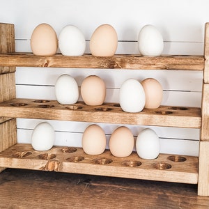 May include: A three-tiered wooden egg holder with a rustic finish. The holder is filled with a mix of white and light brown eggs. The wooden structure has a natural, aged appearance, with visible wood grain.