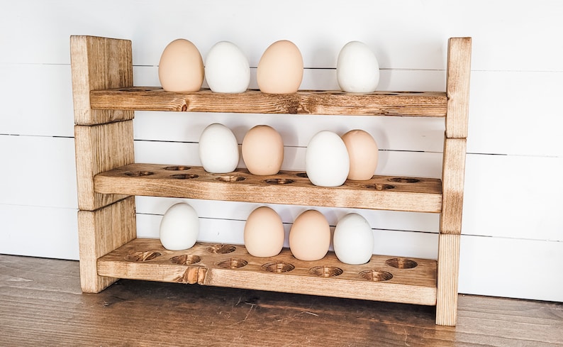 May include: A three-tiered wooden egg holder with a rustic finish. The holder is filled with a mix of white and light brown eggs. The wooden structure has a natural, aged appearance, with visible wood grain.