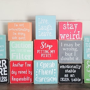 Puede incluir: Una colección de bloques de madera coloridos con frases humorísticas en letras blancas. Los bloques son de varios colores, incluyendo azul, rosa y verde. Las frases incluyen "Stay Weird" y "Purr More Hiss Less."
