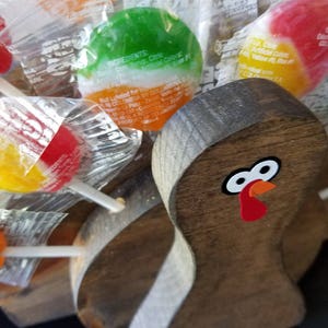 May include: A wooden turkey figurine with a red, orange, and white painted face. The turkey is surrounded by colorful lollipops wrapped in clear plastic.