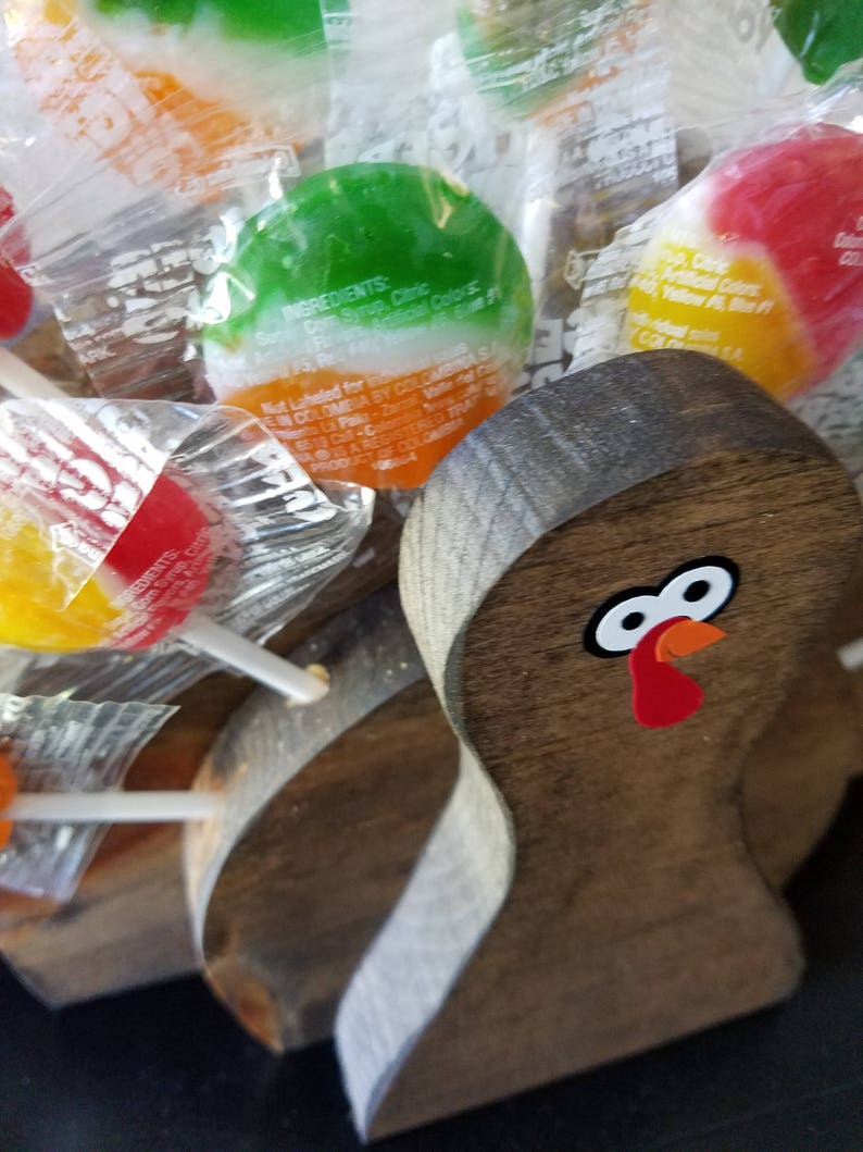 May include: A wooden turkey figurine with a red, orange, and white painted face. The turkey is surrounded by colorful lollipops wrapped in clear plastic.