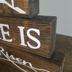 May include: Wooden sign with the words "He is Risen" in white lettering. The sign is made of three stacked pieces of wood.