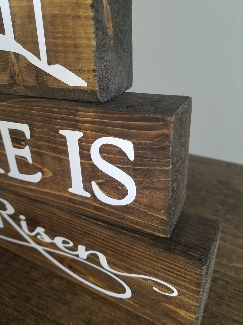 May include: Wooden sign with the words "He is Risen" in white lettering. The sign is made of three stacked pieces of wood.
