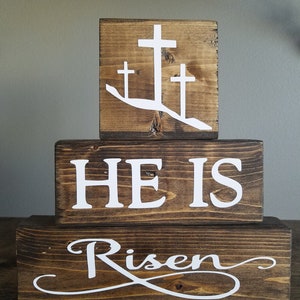 May include: Three wooden blocks stacked on top of each other. The top block has a white silhouette of three crosses on a brown background. The middle block has the words "HE IS" in white letters on a brown background. The bottom block has the word "Risen" in white cursive letters on a brown background.