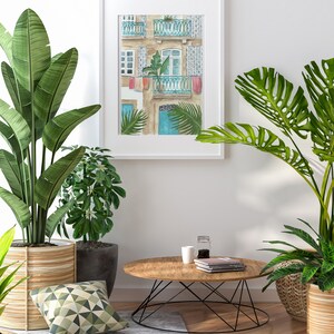 May include: A watercolour painting of a building with a blue door and balconies, framed in white and hanging on a white wall.  Two potted plants, one with green leaves and one with large, green leaves, are on either side of the painting. A wooden coffee table with a black metal base sits in front of the painting.  A white mug and a glass jar with a lid are on the table.