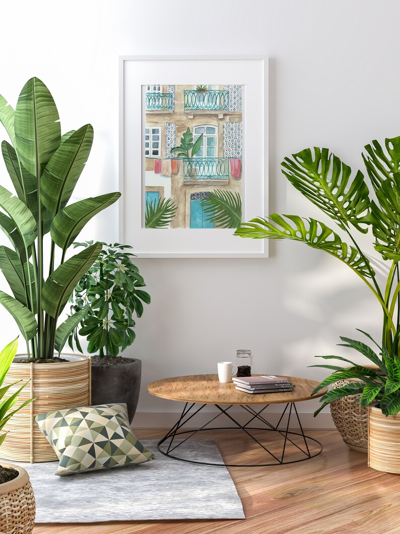 May include: A watercolour painting of a building with a blue door and balconies, framed in white and hanging on a white wall.  Two potted plants, one with green leaves and one with large, green leaves, are on either side of the painting. A wooden coffee table with a black metal base sits in front of the painting.  A white mug and a glass jar with a lid are on the table.