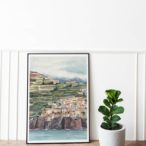 May include: A watercolor painting of a coastal town nestled on a hillside with terraced gardens. The town is painted in shades of yellow, orange, and red, with a blue sea in the foreground. The painting is framed in black and displayed on a white wall.