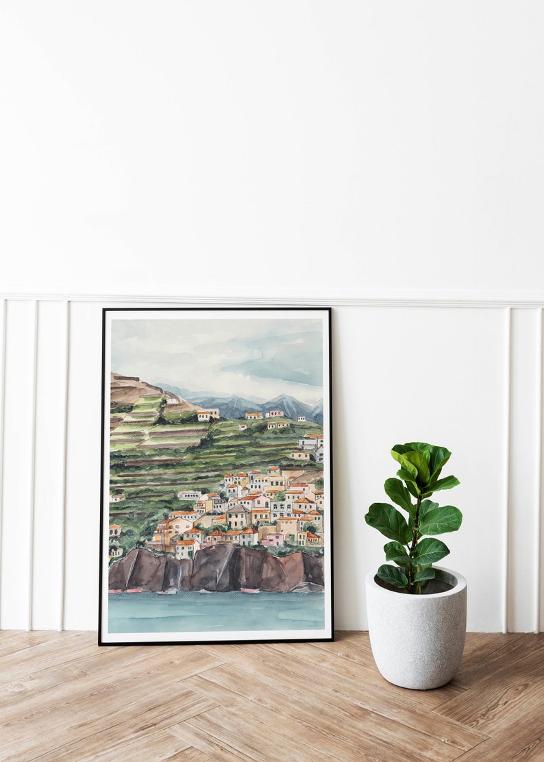 May include: A watercolor painting of a coastal town nestled on a hillside with terraced gardens. The town is painted in shades of yellow, orange, and red, with a blue sea in the foreground. The painting is framed in black and displayed on a white wall.