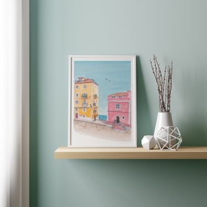 May include: A framed watercolor painting of two buildings on a beach. The building on the left is yellow with white trim and the building on the right is pink with a white door. The sky is blue with a few white clouds.