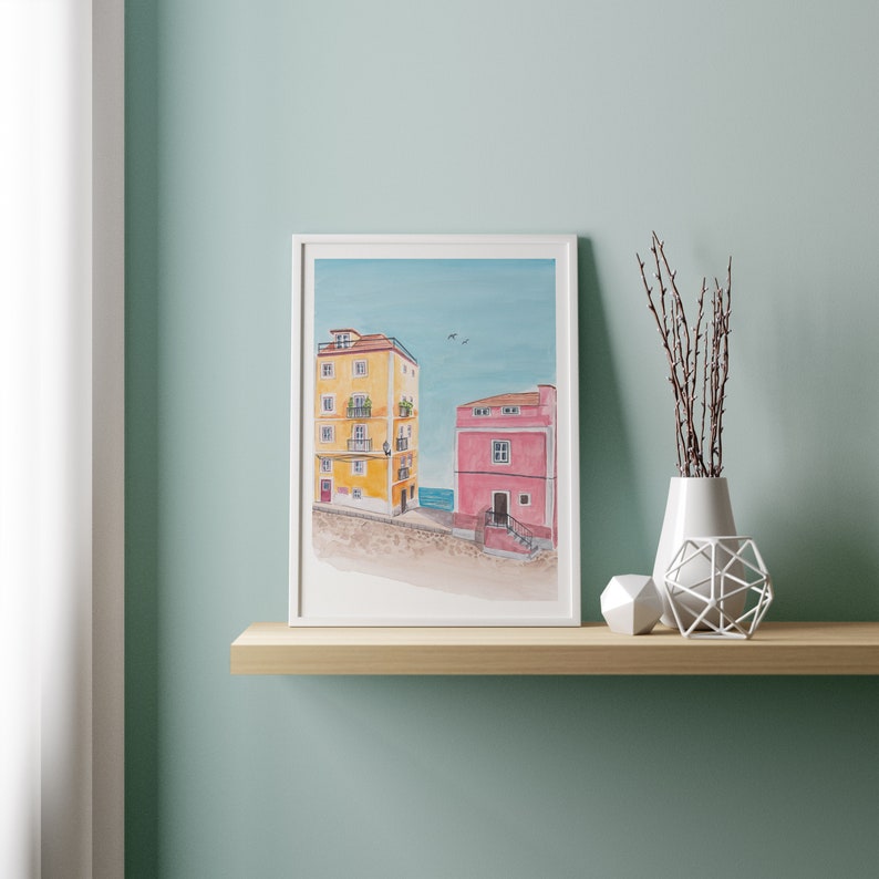 May include: A framed watercolor painting of two buildings on a beach. The building on the left is yellow with white trim and the building on the right is pink with a white door. The sky is blue with a few white clouds.