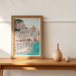 May include: A watercolor painting of a coastal town with colorful buildings, a stone archway, and a beach with blue and white striped umbrellas. The painting is framed in a light brown wood frame.