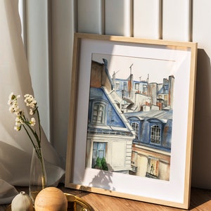 Puede incluir: Una pintura de acuarela enmarcada de un paisaje urbano parisino con edificios y tejados azules y blancos. La pintura est&aacute; en un marco de madera claro y est&aacute; sobre un suelo de madera.