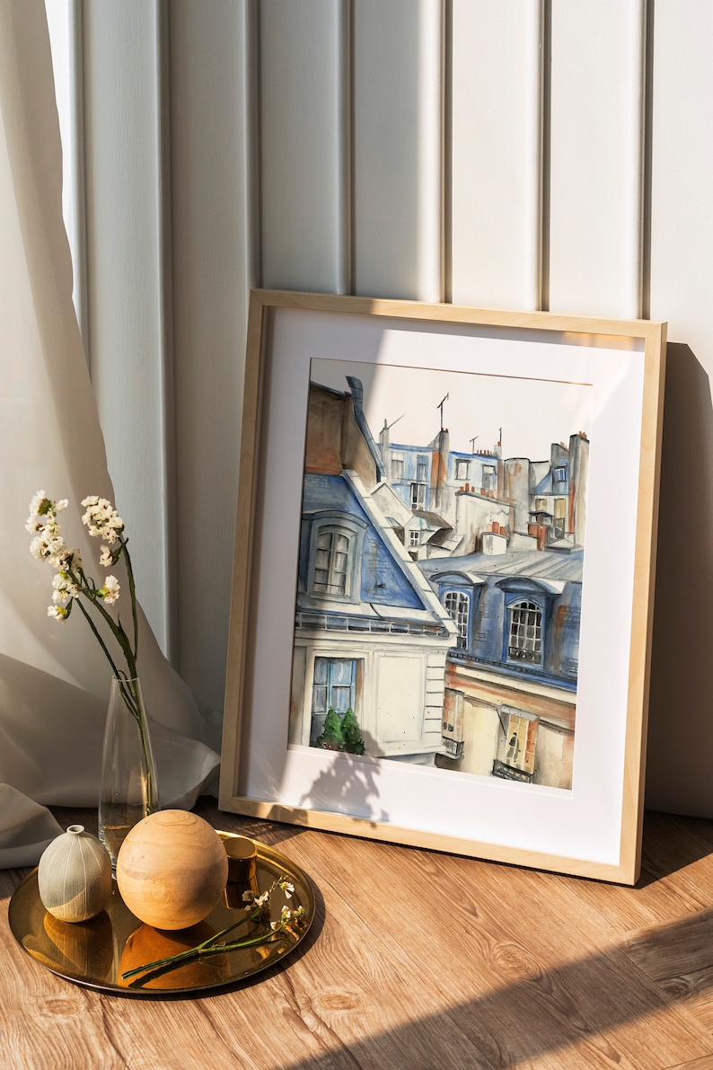 Puede incluir: Una pintura de acuarela enmarcada de un paisaje urbano parisino con edificios y tejados azules y blancos. La pintura est&aacute; en un marco de madera claro y est&aacute; sobre un suelo de madera.