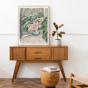 May include: A watercolour painting of a colourful Italian village with a blue car driving up a winding road. The painting is framed in a gold frame and is hanging on a white wall. A wooden side table with two drawers is in front of the painting. A small wooden stool with a stack of books on top is in front of the table. A potted plant is on the table to the right of the painting.