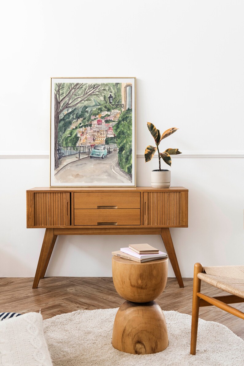May include: A watercolour painting of a colourful Italian village with a blue car driving up a winding road. The painting is framed in a gold frame and is hanging on a white wall. A wooden side table with two drawers is in front of the painting. A small wooden stool with a stack of books on top is in front of the table. A potted plant is on the table to the right of the painting.